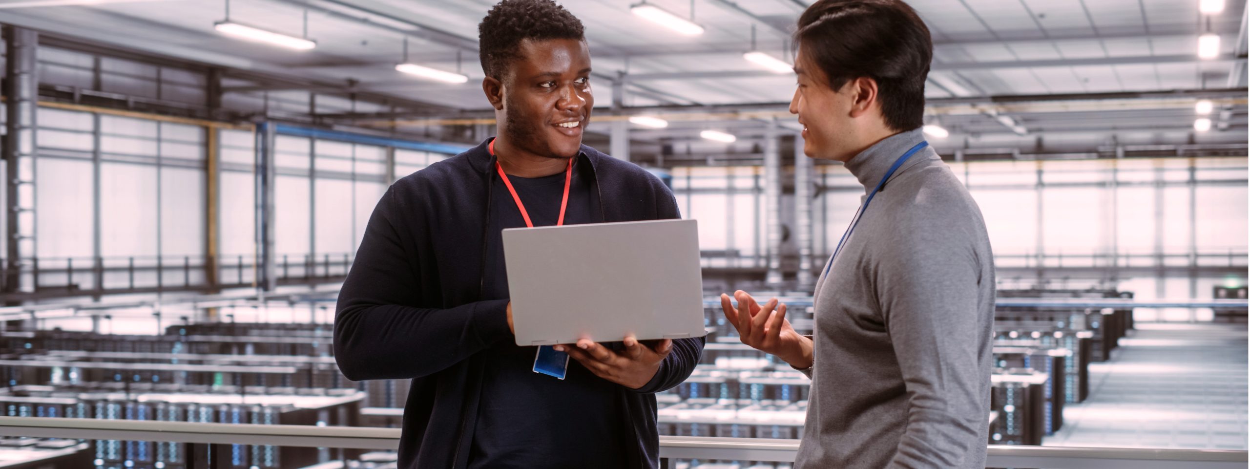 Two Multiethnic Male Data Center System Administrators and IT Specialists Talk, Use Laptop. Information Technology Engineers work on Cyber Security Network Protection in Cloud Computing Server Farm.